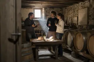 Artisan winemaker hosting a couple in a stone cellar with wine barrels, natural light and rustic ambiance, evoking intimate discovery in the Pays de Caux.