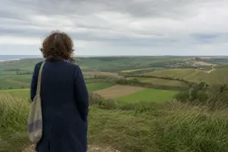 Solitary woman in natural attire thoughtfully observing the rolling Caux plateau and distant vineyards from a windy crest path, under soft Norman sky