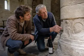 Winemaker and young adult share a subtle moment in a Romanesque church nave, softly lit, with a bottle of Mondeuse wine at their feet, seen from a close and immersive angle.