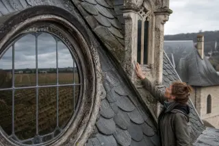 Woman quietly exploring an old wine estate interior, touching a turret window, with vineyards reflected in a lucarne under soft daylight.