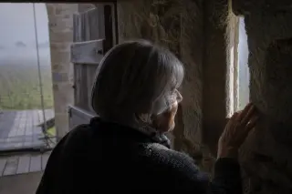 Elderly woman examining an arrow slit inside a Norman wine fortification, soft sunlight streaming through onto stone walls, hint of a drawbridge and misty vineyards in the background.