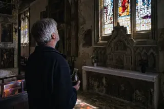 A local winemaker standing inside a gothic church admires colored light from stained glass, holding a carafe of wine, with soft Norman daylight illuminating the ancient stone interior.
