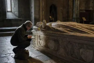 Elderly historian kneeling beside a carved gisant in a softly lit Gothic abbey, bathed in gentle natural light.