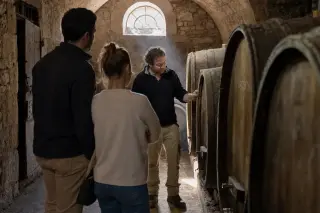 In a medieval stone wine cellar, a winemaker discusses barrels with a young couple, soft daylight streaming in and dust motes visible in the air.