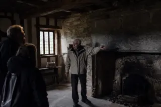 Elderly guide narrating in the dim interior of a Norman manor, light filtering through a small window onto historic wood beams and stone, creating an authentic, contemplative mood.
