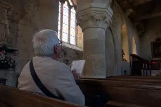 Interior of a Norman church where an elderly local sketches a stone capital, natural daylight filtering through stained glass, quiet and contemplative atmosphere.
