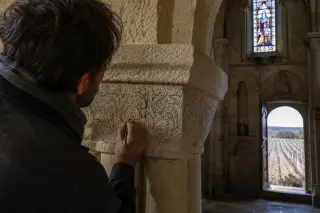 Artisan seen from behind sketching details on a medieval chapiteau inside a Romanesque church, soft daylight highlighting the ancient stone and subtle church interior details.