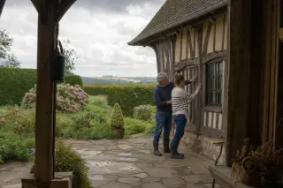 A Norman farmer and his daughter examine wooden barn details in a rustic courtyard, surrounded by lush hedges and stone, under overcast skies.